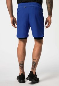 Shorts de sport bleus avec une taille élastique et une poche arrière. La personne porte des shorts de compression noirs et des baskets noires.
