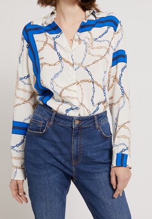 Frau trägt eine weiße Bluse mit blauen und beigen Kettenmustern, die in eine hochgeschnittene blaue Jeans gesteckt ist.