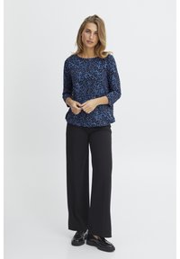 Blusa a maniche tre quarti blu e nera con motivo, abbinata a pantaloni larghi neri e scarpe nere, in piedi di fronte a uno sfondo neutro.