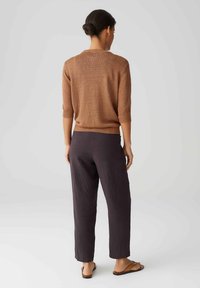 Kobieta z włosami w kokach, ubrana w brązowy sweter z rękawami 3/4, ciemne proste spodnie i beżowe sandały, stojąca na jednolitym szarym tle.