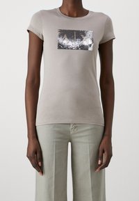 Camiseta de manga corta beige con un parche rectangular de lentejuelas plateadas en la parte delantera. Material de algodón suave con un diseño ajustado.
