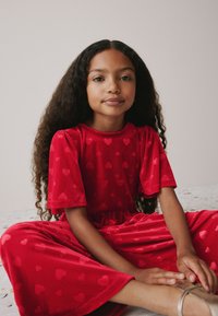 Completo in velluto rosso con maniche corte, caratterizzato da un motivo a cuori su tutta la superficie. Il design presenta una vita arricciata e pantaloni a gamba larga.