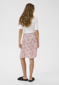 Weißes, tailliertes Oberteil mit kurzen Ärmeln, kombiniert mit einem rosa Rock im Blumenmuster; getragen mit schwarzen Rutsch-Sandalen. Die Haare sind wellig und locker.