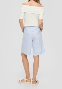 Femme debout de dos portant un haut blanc décolleté, un short rayé bleu à hauteur du genou et des sandales beige.