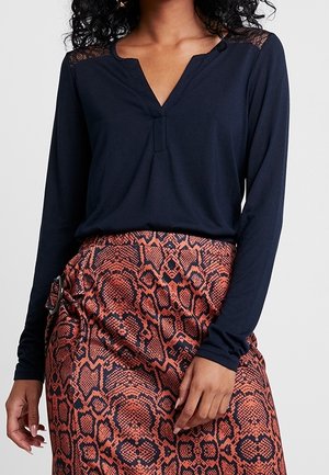 Kobieta ubrana w czarną bluzkę z długim rękawem i dekoltem w serek oraz spódnicę z wysokim stanem w czerwono-czarny wzór skóry węża z bocznym detalem w postaci klamry.