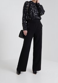Svart långärmad blus med vit stjärntryck, tillsammans med svarta högmidjade vida byxor med dekorativa sidoknappar och en slät textur.