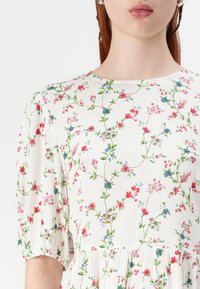 Abito a stampa floreale con maniche a sbuffo, scollatura tonda e un tessuto bianco testurizzato con dettagli floreali colorati in rosso, blu e verde.