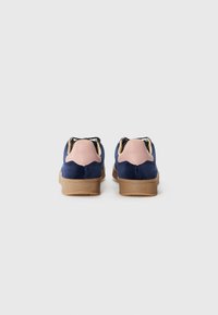 Marineblå suedingsko med en rosa skinnbak, svarte snører og beige gummisåle. Har et bølget design på hælområdet.