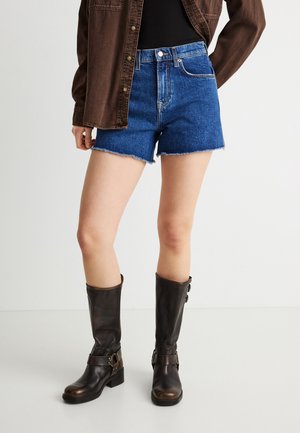 Person trägt blaue Jeansshorts mit ausgefransten Säumen, ein dunkelbraunes Hemd mit Knöpfen und kniehohe dunkelbraune Lederstiefel.