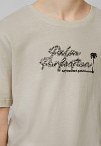 Beżowa bawełniana koszulka z krótkim rękawem, z czarnym napisem "Palm Perfection" i motywem palmy. Prosta, gładka tekstura.