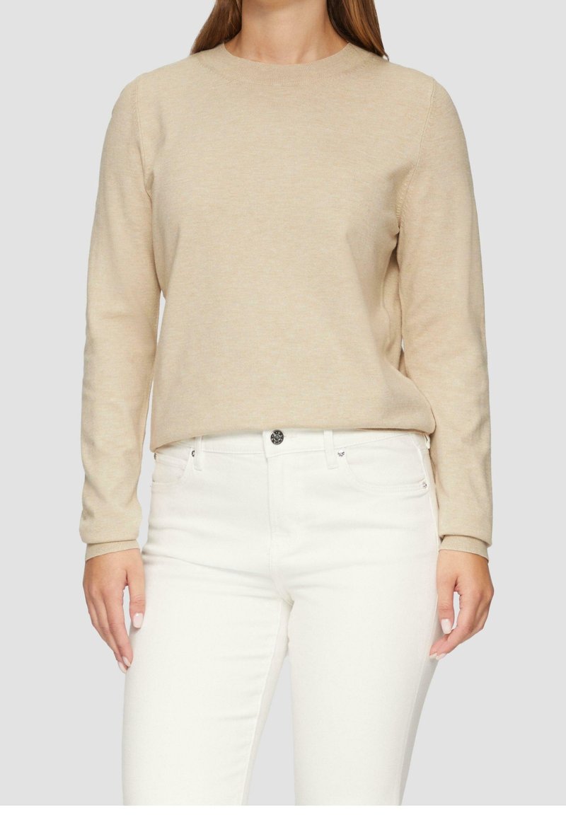 Pull beige à manches longues avec encolure ronde, texture douce et poignets côtelés, associé à un jean blanc. Design simple et épuré.