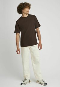 T-shirt marron à manches courtes et côtes associé à un pantalon larges couleur crème. Le mannequin se tient devant un fond neutre et porte des baskets décontractées.