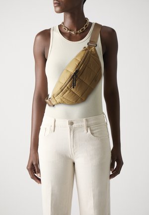 Quilted brown sling bag with zippered pockets, worn over a light tank top. Model wears cream high-waisted jeans and a gold chain necklace.