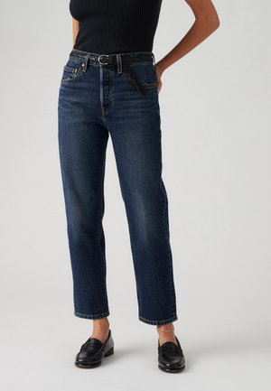 Kvinde iført mørkeblå jeans med lige ben med sort vævet bælte, sort ærmeløs top, sorte loafers og en ankelkæde på højre ankel.