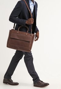 Brun läder budväska med dubbla handtag och en axelrem, med en framficka och slät yta, bärs av en stående figur.