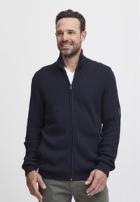 Námornícky pletený sveter na zips s vysokým golierom, z ribovaného materiálu a logom na ľavom rukáve. Obsahuje dlhé rukávy a priliehavý strih.