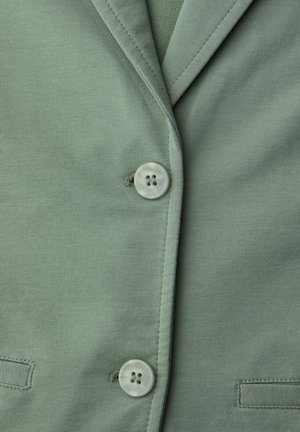 Gros plan sur le devant d'une veste vert clair avec deux boutons et des poches cousues sur un tissu doux.