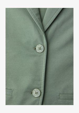 Zbliżenie na przód jasnzie zielonej kurtki z dwoma guzikami i przeszywanymi kieszeniami na miękkiej tkaninie.