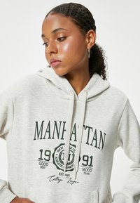 Sudadera con capucha de color gris claro que presenta un estampado gráfico en verde oscuro que incluye "MANHATTAN", números y texto en cursiva. Cuerda de ajuste y bolsillo.