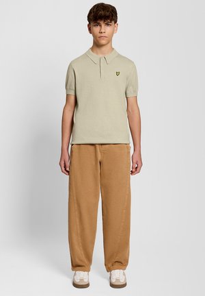 Teenager wearing beige short-sleeve polo with yellow logo, loose brown pants, and white sneakers standing against plain light background.