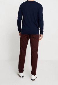 Man i marinblå långärmad skjorta, mörkröda byxor och vita sneakers som står vänd bort från kameran mot en enfärgad vit bakgrund.