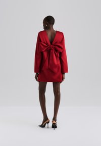 Red satin dress with long sleeves, deep V-back, and oversized bow detail at the waist. Minimalist design with a short hemline.