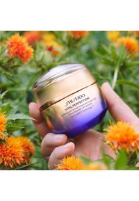 Shiseido Vital Perfection -voide pyöreässä lasipurkissa, jossa kullankeltainen siniseen liukuvärjätty kansi ja tuotetiedot. Puutarhatausta kukkien kanssa.