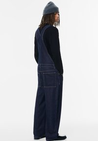 Temne modri jeans overall z X-nazaj obliko, ki ima dva velika sprednja žepa in široke odprtine za noge, oblečeni čez črno majico z dolgimi rokavi.