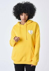 Sudadera amarilla en mezcla de algodón, con un bolsillo frontal, capucha con cordón y un gráfico de un corazón roto en blanco en el pecho.