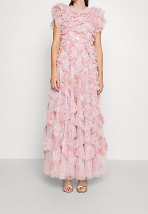 Kobieta ubrana w długą do ziemi, bezrękawną różową suknię z falbankami i kwiatowymi wzorami oraz srebrne sandały na otwartym obcasie, stojąca na tle jednolitego tła.