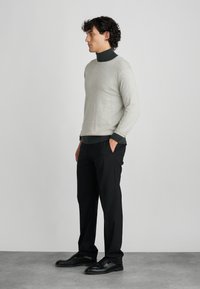 Gai pelēks džemperis ar apaļo kakla izgriezumu, virs tumša augsta kakla krekla, saskaņots ar melniem biksēm un melniem apaviem. Gludas tekstūras.