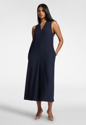 Femme debout portant une combinaison sans manches bleu marine à jambes larges et des chaussures à talons hauts dorées, les mains dans les poches, sur un fond blanc uni.