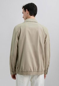 Chaqueta ligera beige con un corte relajado, dobladillo elástico y puños abotonados. El material parece suave con un ligero brillo.