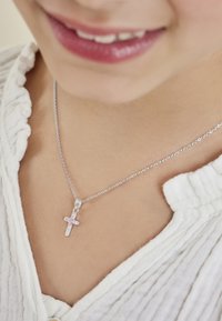 Silvrig korshalsband med en rosa ädelsten, prytt med en delikat kedja. Korset har en strukturerad design och bärs nära en vit, volangblus.