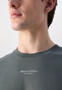 Mørkegrønn bomull T-skjorte med rund hals. Har hvit logo tekst "Marc O'Polo EST. I STOCKHOLM" på forsiden. Glatt tekstur.