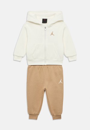 Witte zip-up capuchonjas met zakken aan de voorkant en beige joggingbroek. Beide hebben het Jordan-logo in contrasterende kleuren. Zachte stoftextuur.