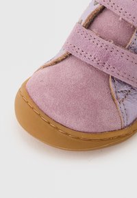 Zapato de ante rosa con acentos de cuero morado, con punta redonda y dos correas ajustables de Velcro. Suela de goma de color marrón claro.