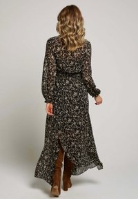 Femme aux cheveux blonds ondulés portant une robe maxi florale noire et des bottes en cuir suédé marron montantes, marchant de dos.