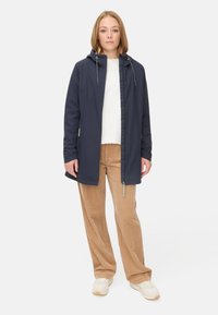 Veste imperméable bleu marine avec capuche, cordon de serrage et deux poches latérales, associée à un pantalon en velours côtelé beige et des baskets beiges.