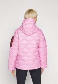 Roze pufferjas met capuchon, voorzien van een golvend quiltpatroon en bordeauxrode accenten op de linker mouw. Lichtgewicht stof.