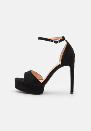 Sort platform sandal med høj hæl, ankelrem og åben tå, vist fra siden på en ensfarvet lys baggrund.