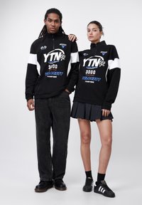 Black zip-up sweatshirt with white accents and blue graphics, paired with dark jeans and a black pleated skirt. Black shoes complete the look.