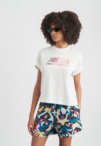 Λευκό κοντομάνικο T-shirt με το γράφημα "new balance", συνδυασμένο με πολύχρωμα σορτς με αφηρημένο σχέδιο. Το μοντέλο φοράει μαύρα γυαλιά ηλίου.