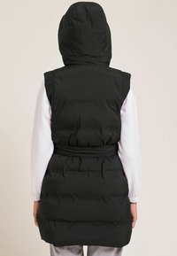 Czarna pikowana kamizelka z kapturem, przewiązana paskiem w talii, o pikowanej fakturze. Tył ma gładkie, czyste wykończenie.