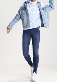 Chaqueta de mezclilla, sudadera ligera azul con texto en negrita, jeans ajustados oscuros, zapatillas blancas; atuendo casual con detalles de mezclilla desgastada.