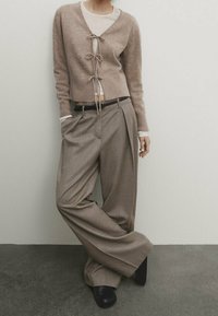 Beige värinen lyhyt neuletakki, jossa on solmittavat nauhat, kerrostettuna vaalean topin päälle, yhdistettynä leveälahkeisiin harmaisiin ruutuihin ja mustiin korokepohjakenkiin.