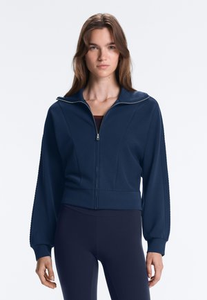 Junge Frau mit langen braunen Haaren trägt eine marineblaue Jacke mit Reißverschluss und passende Leggings, steht vor einem schlichten hellen Hintergrund.