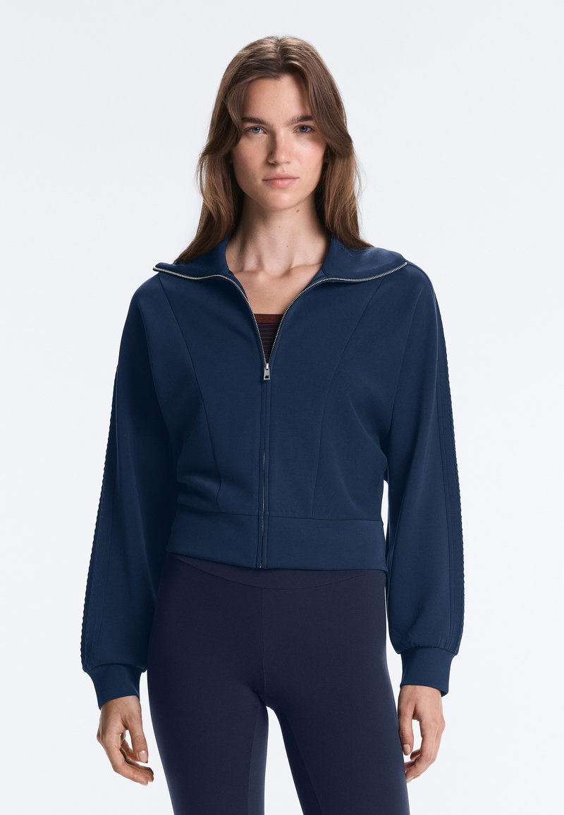 Jeune femme aux longs cheveux bruns porte une veste zippée bleu marine et des leggings assortis, debout devant un fond clair uni.