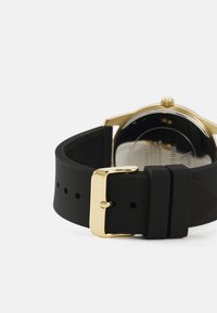 Black rubber watch strap with a gold metal buckle. The watch face is partially visible, showcasing a clear glass cover and gold accents.