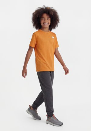 Χαμογελαστό παιδί με κατσαρά μαλλιά που περπατά, φορώντας πορτοκαλί μπλουζάκι, σκούρο γκρι παντελόνι και γκρι αθλητικά παπούτσια σε λευκό φόντο.